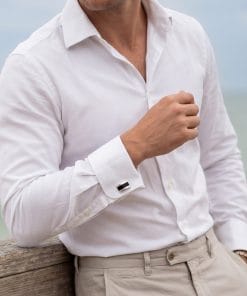 Man in a white button-down shirt and beige pants resting his left arm on a weathered wooden railing outdoors. The scene focuses on the torso and cuff with a small cufflink visible on the sleeve.